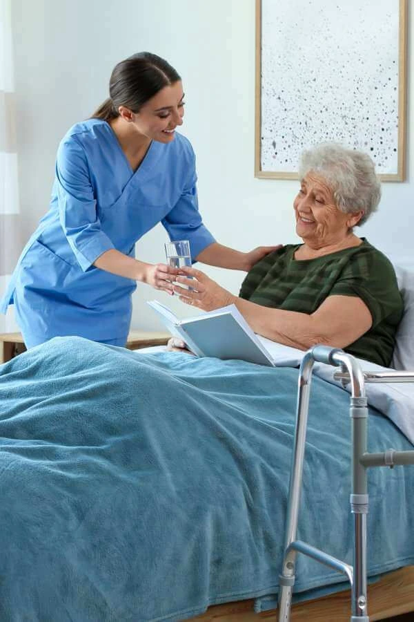 A female caregiver giving a glass of water to an elderly woman laying on the bed
